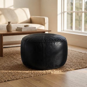 May include: A round, black leather ottoman featuring a smooth, matte finish with visible vertical stitched seams that divide the surface into panels. The piece has a low-profile, cylindrical shape with a flat top and a structured, firm appearance.