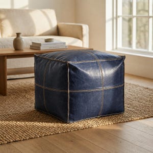 Pode incluir: Um pufe quadrado de couro azul-marinho profundo com costuras de contraste claras e proeminentes que formam um padrão de grade em suas superfícies. O apoio para os pés em forma de cubo tem uma textura lisa, levemente desgastada e bordas definidas.