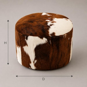 May include: A round ottoman upholstered in brown and white cowhide with a short-haired texture. The cylindrical piece features a distinct spotted pattern across the top and sides. Dimension lines labeled H and D are visible on the sides.
