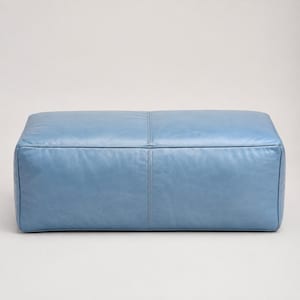 Pode incluir: Um pufe retangular estofado em couro liso azul-claro. A peça apresenta um design limpo e estruturado com uma costura vertical proeminente que percorre o centro da superfície superior e da face frontal, criando uma aparência equilibrada.