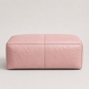 May include: A rectangular ottoman upholstered in a soft, dusty pink leather material. The piece features a clean, structured design with a prominent vertical seam running down the centre of the front panel and visible piping along the top edges.