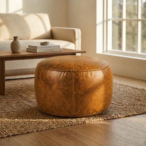 Könnte beinhalten: Ein runder Pouf-Hocker aus beigem Leder mit einer strukturierten, abgenutzten Oberfläche und sichtbaren vertikalen Nähten. Das zylindrische Möbelstück hat eine flache Oberseite und eine feste, strukturierte Form mit einer natürlichen, gebrauchten Optik.