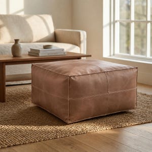 Pode incluir: Um otomano quadrado de couro na cor castanho-claro com costuras visíveis que dividem o topo e as laterais em painéis. O otomano possui um acabamento liso e fosco com um design estruturado de perfil baixo, apoiado sobre um tapete tecido.