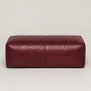 May include: A rectangular ottoman upholstered in deep burgundy leather. The piece features a smooth, continuous surface with a single vertical seam running down the center of the top panel and matching seams along the front face, creating a structured look.