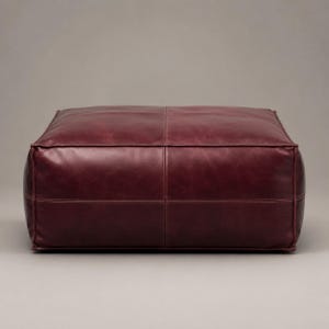 May include: A square, low-profile floor cushion upholstered in deep burgundy leather. The surface features a smooth texture with visible stitched seams forming a cross pattern across the top and sides, creating a structured, boxy silhouette.