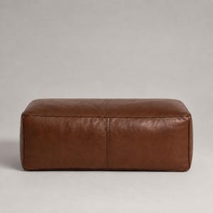 May include: A rectangular ottoman upholstered in smooth, medium-brown leather. The piece features a clean, boxy silhouette with a central vertical seam running across the top and down the front face, showcasing a consistent, uniform leather texture.