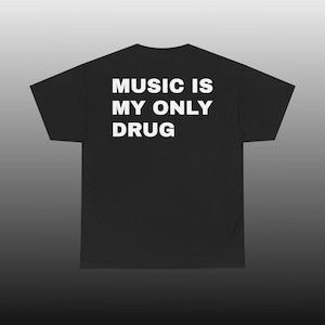 Può includere: Una t-shirt nera a maniche corte con il testo bianco "MUSIC IS MY ONLY DRUG" stampato in un carattere sans-serif in grassetto sulla parte superiore della schiena. Il capo presenta un girocollo standard e un orlo dritto, mostrato in piano.