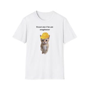 Può includere: Una t-shirt bianca a maniche corte con la grafica di un piccolo gattino arancione che indossa un casco da cantiere giallo. Sopra il gattino, il testo Trust me i'm an engineer è scritto in caratteri neri senza grazie.