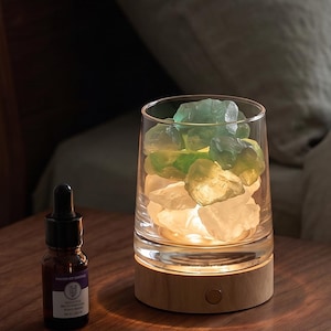 May include: A cylindrical glass vessel filled with translucent green and white stones, illuminated from a light-colored wooden base. Beside the lamp sits a small dark brown glass dropper bottle with a white label and a black dropper cap.