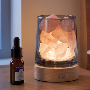 May include: A cylindrical glass lamp filled with rough, translucent pink salt rocks, set on a circular light-toned wooden base with a metallic power button, accompanied by a small brown glass dropper bottle with a white label and black cap.