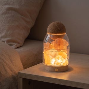 May include: A glass jar lamp with a cork lid containing glowing orange salt crystals. The glass features the printed text: I SEE THE MOUNTAINS RIVERS THE SUN MOON AND THE STARS WHERE MY EYESIGHT IS WHERE YOU ARE. It has a wooden base.