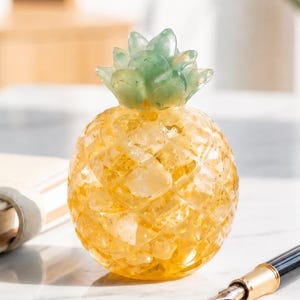 Puede incluir: Un objeto decorativo con forma de piña elaborado con material cristalino amarillo y verde. El cuerpo presenta una superficie texturizada y facetada que recuerda a la piel de la piña, rematada con un grupo de hojas lisas de color verde claro.