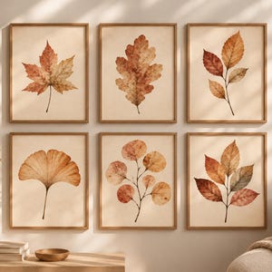 Könnte beinhalten: Ein Set aus sechs gerahmten Wandbildern mit Aquarellillustrationen verschiedener Herbstblätter in Orange-, Braun- und Rottönen. Jeder Druck ist in einem hellen Holzrahmen gefasst und zeigt ein einzelnes Blatt oder einen Zweig auf cremefarbenem Grund.