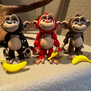 May include: Three articulated monkey figurines in black, red, and brown. Each monkey features a tan face, chest, and hands, with large eyes and a smiling expression. Each figure is accompanied by a small, bright yellow plastic banana accessory.