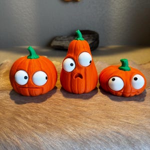 May include: Three orange pumpkins with green stems and large, white, googly-style eyes. The left pumpkin is round, the center pumpkin is tall and pear-shaped with a surprised expression, and the right pumpkin is short and wide.