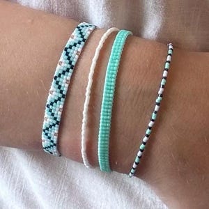 Könnte beinhalten: Ein Set aus vier Perlenarmbändern. Eines zeigt ein geometrisches Zickzackmuster in Hellblau, Weiß und Schwarz. Ein weiteres besteht aus einer einzelnen Reihe weißer Perlen. Das dritte ist ein einfarbiges, hellblau gewebtes Band. Das vierte ist ein dünnes Armband aus abwechselnd schwarzen, weißen und hellblauen Perlen.