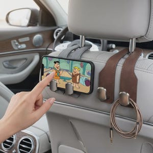 May include: A set of two car headrest hooks made of leather-like material in grey and brown, featuring integrated metal clips. The grey hook holds a smartphone displaying a cartoon, while the brown hook holds a coiled charging cable.