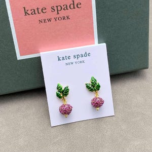 Puede incluir: Un par de pendientes colgantes Kate Spade New York con un tallo de metal dorado, hojas de esmalte verde y un dije redondo de verdura incrustado con cristales rosas, presentados en una tarjeta blanca con el texto Kate Spade New York.