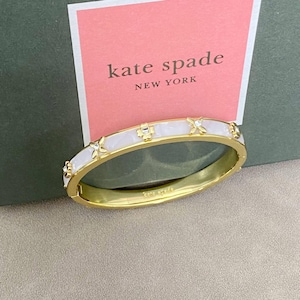 Könnte beinhalten: Ein goldfarbener Klapparmreif mit weißer Emaileinlage und kleinen, blumenförmigen Metallakzenten, die mit klaren Kristallen besetzt sind. Die Innenseite des Armreifs ist mit dem Schriftzug kate spade graviert, platziert vor einer rosa Karte.