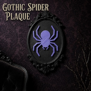 May include: An oval black frame with ornate detailing containing a solid purple spider silhouette. The text GOTHIC SPIDER PLAQUE is printed in the upper left corner. The spider has a round body and eight angular legs centered on a black background.