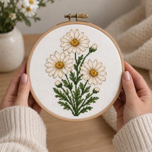 Puede incluir: Un bastidor de bordado circular de madera que contiene tela blanca con un patrón de punto de cruz de tres margaritas blancas con centros amarillos y tallos y hojas verdes. El diseño presenta varios capullos pequeños sin abrir.