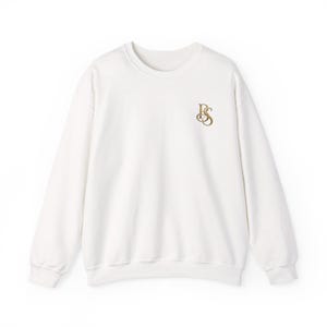 May include: A white long-sleeve crewneck sweatshirt featuring a gold-colored embroidered monogram logo on the upper left chest. The logo consists of the intertwined letters RS in a serif font. The garment has ribbed cuffs and a ribbed hem.