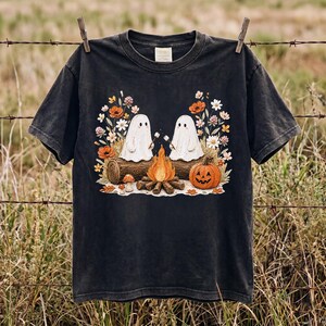 Puede incluir: Una camiseta negra de manga corta con un estampado gráfico de dos fantasmas blancos sentados en un tronco junto a una hoguera, asando nubes. El diseño incluye una calabaza naranja tallada y varias flores silvestres de colores.