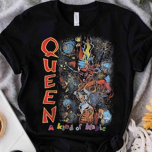 Puede incluir: Una camiseta negra de manga corta con un estampado gráfico que muestra la palabra QUEEN en letras rojas grandes verticalmente en el lado izquierdo. El diseño incluye una ilustración abstracta y colorida de una figura y el texto A kind of Magic.