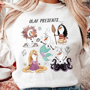 Könnte beinhalten: Ein weißes Sweatshirt mit dem Text OLAF PRESENTS... und einem Grafikdruck der Figur Olaf, verkleidet als verschiedene Gestalten, darunter ein Löwe, eine Meerjungfrau, eine langhaarige Prinzessin, ein Reisender mit Paddel und ein Oktopus.