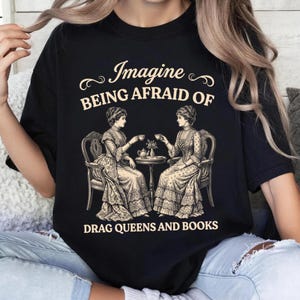 Puede incluir: Una camiseta negra con un gráfico de color crema de dos figuras de estilo victoriano sentadas en una mesa pequeña con tazas de té. El texto "Imagine BEING AFRAID OF DRAG QUEENS AND BOOKS" aparece en una fuente decorativa.