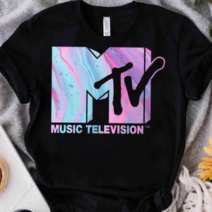 Puede incluir: Una camiseta negra de manga corta con el logotipo de MTV en el pecho. El logotipo está relleno con un patrón jaspeado de remolinos azul claro, rosa y blanco. Debajo del logotipo, el texto MUSIC TELEVISION está impreso en una fuente sans-serif azul claro.