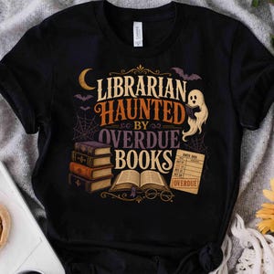 Puede incluir: Una camiseta negra de manga corta con un gráfico que incluye el texto LIBRARIAN HAUNTED BY OVERDUE BOOKS. El diseño muestra un fantasma sosteniendo un libro, una pila de libros, un libro abierto, una tarjeta de biblioteca, una luna, murciélagos y telarañas.