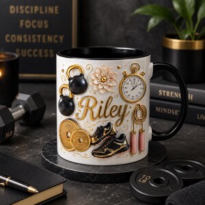 Op de afbeelding: Een witte keramische mok met een zwarte binnenkant en handvat, voorzien van reliëfdecoraties waaronder twee zwarte kettlebells, een gouden stopwatch, een bloemembleem, gouden gewichtsschijven met 10 LB aanduiding, sportschoenen en een springtouw.