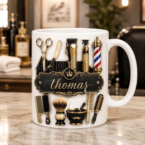 Puede incluir: Una taza de cerámica blanca con un diseño gráfico de herramientas de barbero en dorado y negro, incluyendo tijeras, navaja, cortapelos, poste de barbero, brocha de afeitar y peines. Una placa negra central muestra el nombre Thomas en oro.
