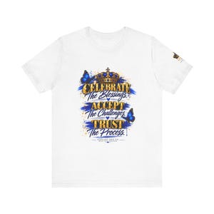 May include: A white short-sleeve t-shirt featuring a graphic print with a gold crown, blue butterflies, and the text: CELEBRATE The Blessings. ACCEPT The Challenges. TRUST The Process. ALREADY HER CO. WEAR YOUR REMINDER. A small logo is on the sleeve.