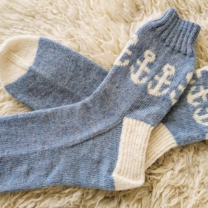Könnte beinhalten: Ein Paar blau-cremefarbene Strickstrümpfe mit einem sich wiederholenden weißen Anker-Muster an den gerippten Bündchen. Die Socken haben einfarbige, cremefarbene Fersen und Zehenbereiche sowie eine melierte blaue Strickstruktur am Hauptteil.
