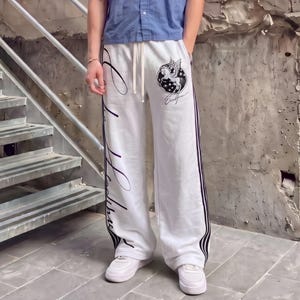 Könnte beinhalten: Weiße Jogginghose mit einer schwarzen Grafik eines geflügelten Pferdes in einem Kreis auf dem linken Oberschenkel, schwarzem Schriftzug vertikal am rechten Bein und schwarzen Seitenstreifen. Die Hose hat einen Kordelzug und weite Passform.