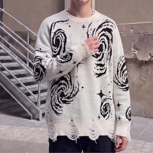 May include: A long-sleeve knit sweater in off-white featuring a black graphic pattern of swirling galaxies and four-pointed stars. The garment has a crew neckline, ribbed cuffs, and a distressed, frayed hemline along the bottom edge.