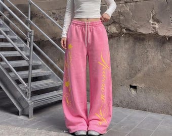 May include: A long-sleeved white crew-neck top paired with bright pink wide-leg sweatpants. The sweatpants feature a high elastic waistband with a white drawstring and yellow graphic prints of stars and cursive text along the legs.