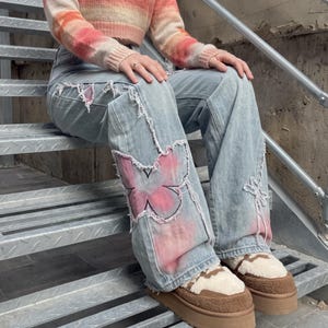 以下が含まれることがあります： 縁がほつれた大きなピンクの蝶のパッチ、ダメージ加工、ワイドレッグフィットが特徴のライトブルーのデニムジーンズ。オレンジ、ピンク、クリーム色のグラデーションのクロップド丈長袖セーターと組み合わされています。