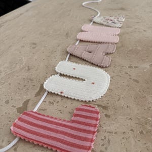 May include: A fabric banner spelling the name LUNA, featuring individual letters strung on a white cord. The letters are crafted from various textiles, including pink stripes, cream with a cable knit texture, solid beige, solid pink, and a floral print.