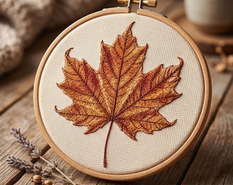 May include: A circular wooden embroidery hoop containing a piece of off-white Aida fabric. Centered on the fabric is a detailed embroidery of a single maple leaf in shades of burnt orange, deep red, and brown, featuring intricate vein stitching.