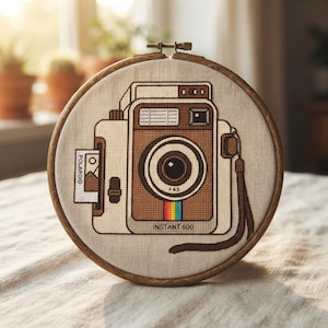May include: A circular wooden embroidery hoop containing a cross-stitch design of a brown and white instant camera. The camera features a lens, a rainbow stripe, a wrist strap, and a small photo ejecting from the side with the text Polaroid.