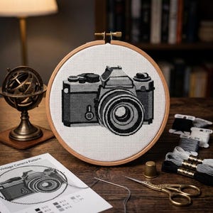 Könnte beinhalten: Ein runder Holzstickrahmen mit einem Kreuzstichmotiv einer Vintage-Kamera in Schwarz- und Grautönen auf weißem Stoff. Die Kamera zeigt ein markantes Objektiv, einen Sucher und mechanische Einstellräder im pixeligen Design.
