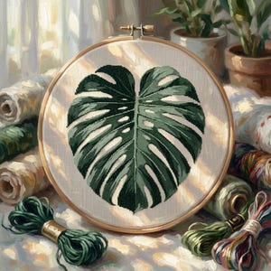 May include: A circular wooden embroidery hoop containing a cross-stitch design of a large, dark green monstera leaf on a light beige fabric background, surrounded by various spools and skeins of embroidery thread in green and earth tones.