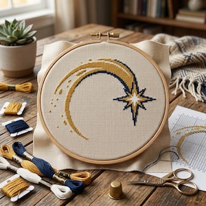 Könnte beinhalten: Ein Kreuzstichprojekt in einem runden Holzstickrahmen mit einem gold- und dunkelblauen Sternschnuppenmotiv auf beigem Aida-Stoff. Das Design zeigt eine geschwungene Spur aus goldenen Stichen, die in einem mehrzackigen Stern mit weißer Mitte endet.