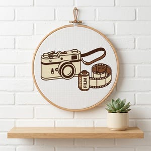 May include: A cross-stitch embroidery in a round wooden hoop featuring a beige and brown vintage camera with a matching strap and a roll of 35mm film with the text 36 EXP FILM printed on the canister, displayed on a white fabric background.