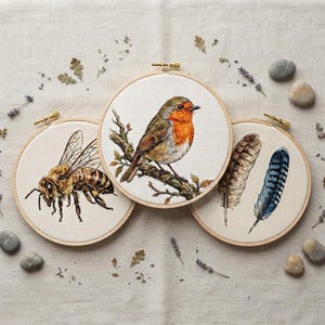May include: Three circular wooden embroidery hoops featuring detailed needlework designs. One hoop displays a honeybee, another shows a robin perched on a branch, and the third features two feathers, one brown and one blue, on cream fabric.
