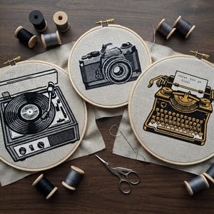 May include: Three circular wooden embroidery hoops containing fabric with black and gold thread designs of a record player, a camera, and a typewriter with paper. Several spools of grey and black thread and metal scissors are scattered around.