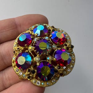 Op de afbeelding: Een ronde goudkleurige broche met een centrale gefacetteerde paars en groen iriserende steen, omringd door vijf grotere bijpassende iriserende stenen. Het metalen frame is gedetailleerd met kleine heldere strass-steentjes en filigraanpatronen.
